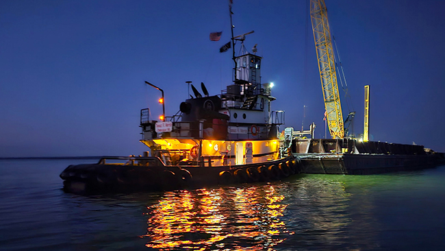 A close-up nighttime photo of a tug and barge. The tug is lit with yellow lights and an American flag is present. The barge has a tall crane that is lit with work lights. The lights on the tug and barge are reflecting on the calm water surrounding both.